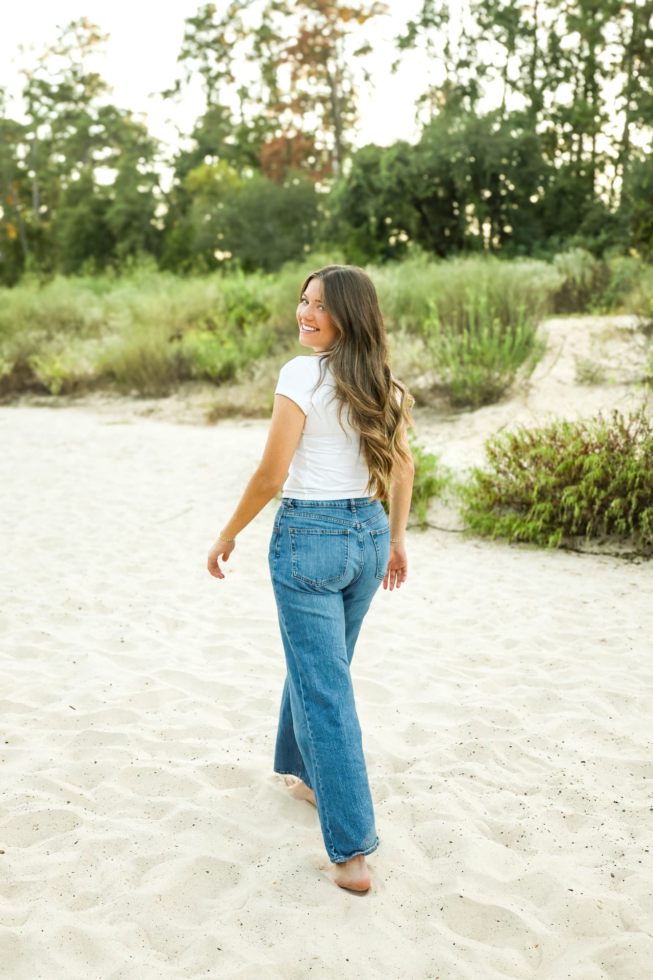 Senior in white tee and jeans walking on sand, looking back over shoulder
